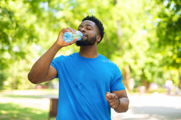Young man drinking water from a plastic bottle while taking a break during outdoor workout. Athlete in blue t-shirt staying hydrated after running in a sunny park.
