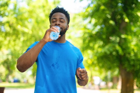 Young man drinking water from a plastic bottle while taking a break during outdoor workout. Athlete in blue t-shirt staying hydrated after running in a sunny park.
