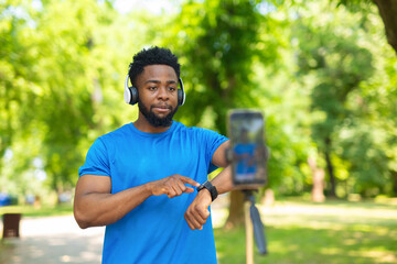 Young african american man showing smartwatch while filming vlog on smartphone tripod outdoors.
