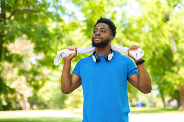 Relaxed young athlete with towel over shoulders and headphones after outdoor training in park.
