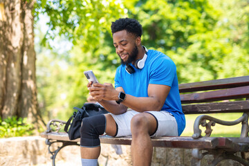 Young man sitting on bench in park, using smartphone after workout.