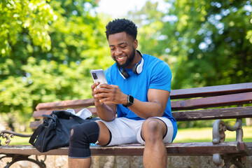 Fototapeta premium Young man sitting on bench with headphones and smartphone smiling outdoors after exercise. 