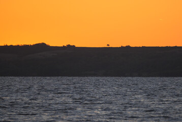 The sun rises from behind the hills. Sunrise on the lake shore. Orange sky.