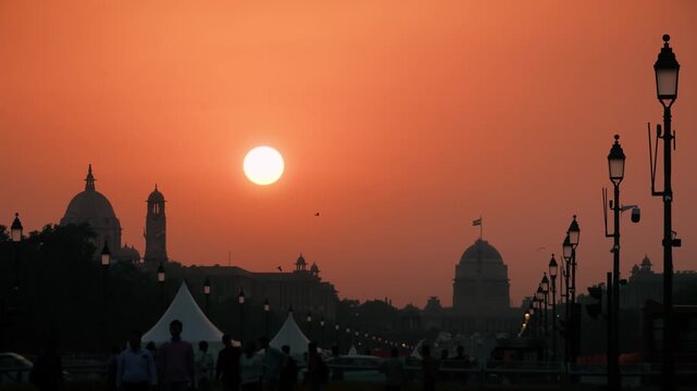 A stunning sunset view of Rashtrapati Bhavan in New Delhi, silhouetted against a deep orange evening sky. The iconic dome and surrounding government buildings appear dramatic in the golden hour.