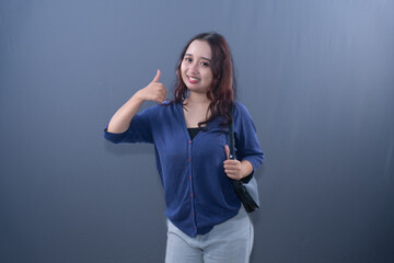 Fototapeta premium Young Asian woman in blue cardigan, smiling, giving thumbs-up, holding bag, grey background