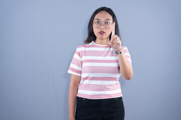 Fototapeta premium Young Asian woman in striped shirt and glasses, pointing finger upwards, serious expression