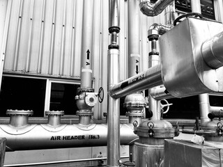 black-and-white image of complex industrial piping and machinery. The detailed composition of valves, tubes, and a corrugated metal wall highlights the intricate and functional aesthetics