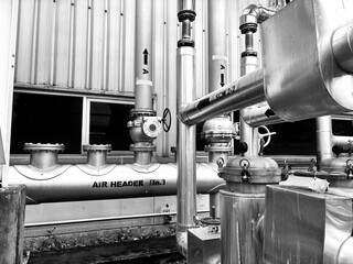 black-and-white image of complex industrial piping and machinery. The detailed composition of valves, tubes, and a corrugated metal wall highlights the intricate and functional aesthetics