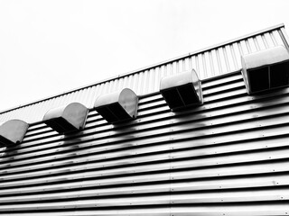 A close-up, low-angle shot of a building's corrugated metal wall, highlighting a series of industrial vents. The black and white photography emphasizes texture and modern, minimalist architecture.