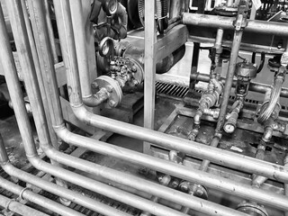 A striking black and white close-up of a complex network of industrial pipes, valves, and gauges. The image focuses on the intricate arrangement of metal tubes and machinery, highlighting the texture