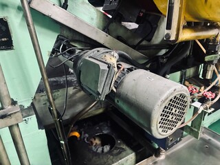 Close-up of a well-worn electric motor on heavy industrial machinery in a factory setting. The image captures the gritty, used texture of the motor and surrounding metal parts, with grease stains,