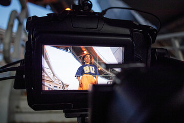 Portrait of girl standing outdoors being filmed by professional camera, looking confidently at lens, modern urban background visible through camera display