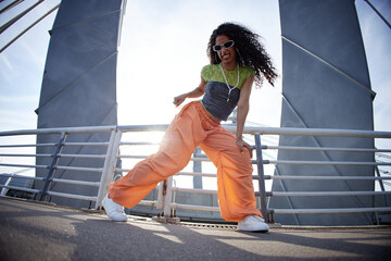 Girl dancing energetically outdoors on urban bridge, wearing sunglasses and headphones, expressing confidence and individuality, sunlight shining behind subject
