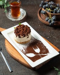 Tiramisu dessert served on a white plate with a chocolate pattern and fresh grapes on dark wooden table