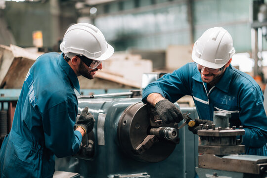 Two factory workers are collaborating on a large piece of industrial machinery. Both men are wearing blue jumpsuits, black work gloves, and white hard hats, with also wearing safety glasses.