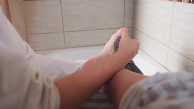 Lower view of schoolgirl seated in bathtub drying feet with soft gray towel, hands wiping heel and sole after shower, focus on hygiene, clean skin, tiled bathroom, relaxed private home routine