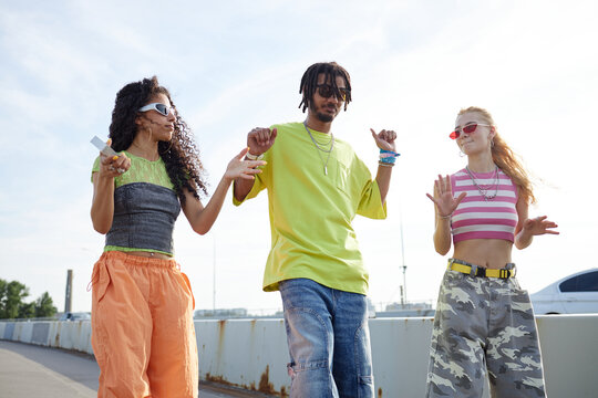 Group of three Gen Z people dancing outdoors on urban street, wearing sunglasses and holding smartphone, expressing carefree attitude - Powered by Adobe