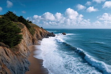 Cliffside ocean view waves crash on a sandy beach below a rocky cliff with green trees under a blue sky with scattered clouds