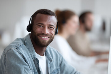 Fototapeta premium A smiling African American man with headset in office. Represents customer service, tech support, communication, teamwork. Perfect for business, online work service needs.