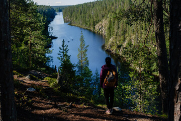 A solitary hiker stands overlooking the serene Canyon Lake Julma Olkky, surrounded by lush forest. The vibrant blue waters reflect the lush greenery, showcasing natures peace in Hossa National Park.