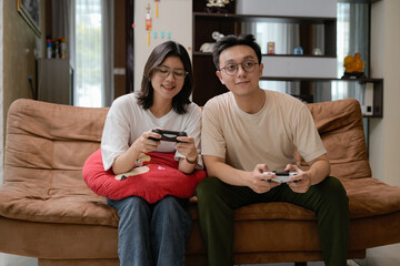 A couple sits close on a brown sofa, smiling while playing video games with handheld controllers