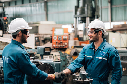 Two male factory workers are shaking hands while standing in a busy workshop. Both men are wearing matching blue jumpsuits, white hard hats, safety glasses, and black gloves. They are smiling