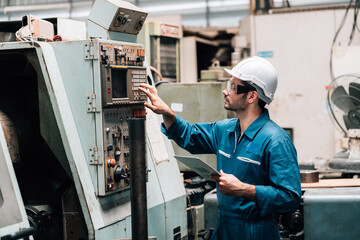 A young male factory worker, wearing a blue jumpsuit, a white hard hat, and safety glasses, is standing in front of a complex piece of industrial machinery. He is holding and looking down at a tablet