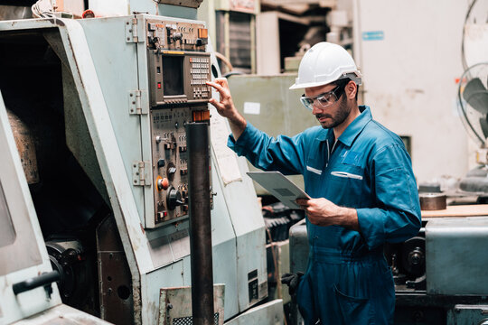 A young male factory worker, wearing a blue jumpsuit, a white hard hat, and safety glasses, is standing in front of a complex piece of industrial machinery. He is holding and looking down at a tablet