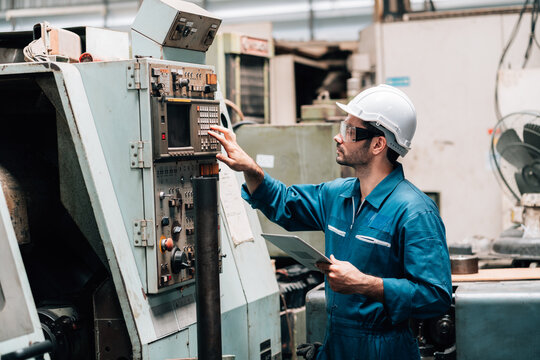 A young male factory worker, wearing a blue jumpsuit, a white hard hat, and safety glasses, is standing in front of a complex piece of industrial machinery. He is holding and looking down at a tablet