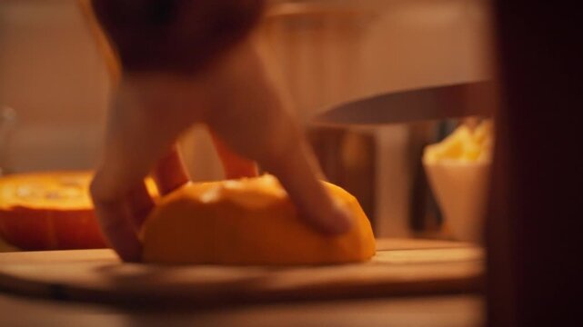 A hand skillfully cuts a piece of bright orange butternut squash on a wooden cutting board in a warm kitchen. The scene captures the essence of cooking during the fall season