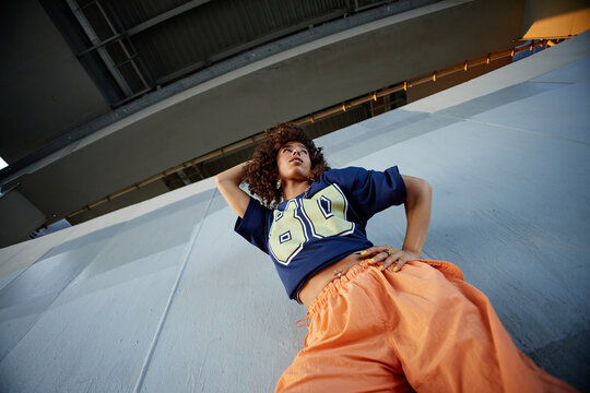 Girl standing outdoors under modern structure, hand on hip and other arm raised behind head, looking away from camera with confident expression, casual streetwear style