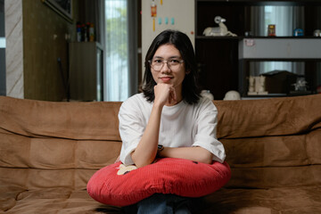 Obraz premium A relaxed woman in a white shirt sits on a brown couch, hugging a red pillow in a cozy living room