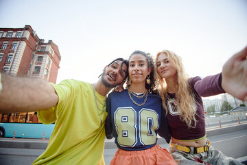 Obraz premium Portrait of three multiethnic peers smiling and taking selfie outdoors in urban setting, two girls and one boy posing closely together, showing friendship and youthful energy