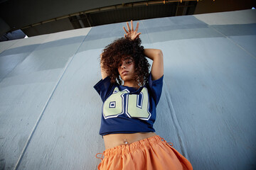 Portrait of girl standing against urban wall, raising arms above head, looking directly at camera, wearing casual clothing, expressing confident attitude
