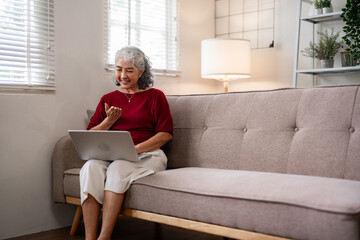 Obraz premium Asian senior woman video calls friends family laptop sofa cozy home connects loved ones digital communication technology lifestyle retirement wellbeing