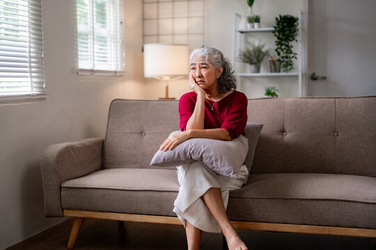 Asian older woman sitting sofa home feeling lonely sad worried contemplating thoughts elderly senior female mental health issue challenges