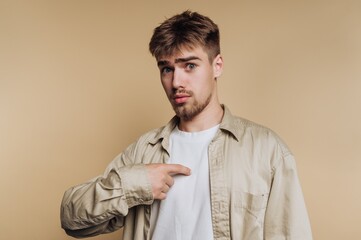 Young man pointing to his chest against a plain background, smiling and engaging in a lighthearted moment during a casual indoor setting