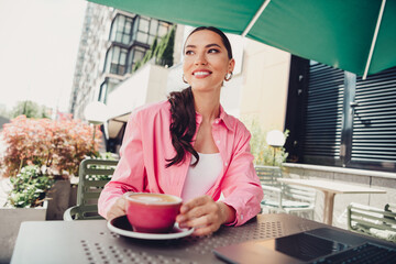 Lovely woman enjoying a coffee in an outdoor urban café on a sunny day, wearing a pink shirt and...