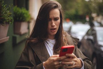 Outdoor shot of serious woman uses modern gadgets walks at street uses mobile phone, wears trench...