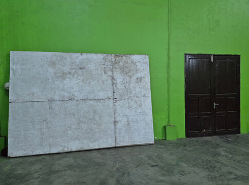 A large, weathered board leaning against a bright green wall next to a closed, dark wooden door on a concrete floor.
