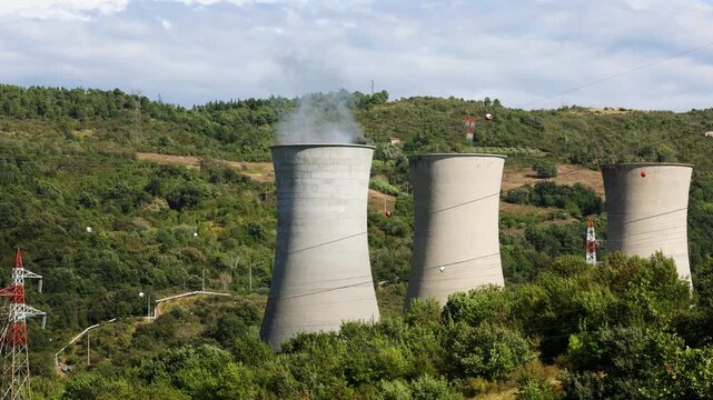 Cooling towers of a thermal power plant release plumes of steam above a green valley under dramatic clouds. Powerful symbol of energy production, air pollution and the green transition, climate change