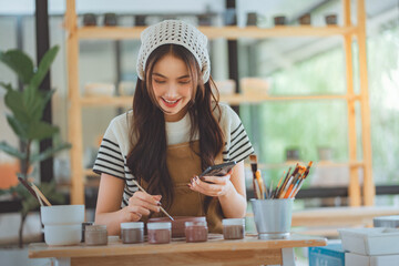 Happy Asian female artist using smartphone to find references and ideas for painting ceramic pottery design in creative studio, woman painting custom artwork for client project