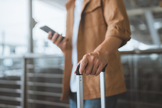 Close-up of Asian male traveler’s hand holding smartphone and suitcase handle at airport terminal – modern travel lifestyle, mobile connectivity, trip preparation journey