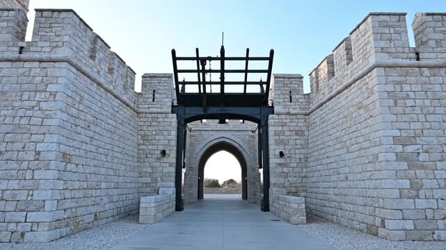 Majestic stone castle entrance with towering crenellated walls and a grand arched gateway featuring a prominent black drawbridge mechanism under a clear sky