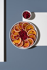 Rodajas de naranja sanguina y semillas de granada en un plato blanco sobre un fondo blanco y azul....