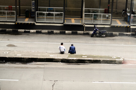 An authentic overhead view captures two men sitting casually on a concrete median in a dynamic urban street, reflecting moments of pause amidst the city's daily bustle and public transport.