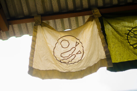 Rustic fabric banner with an abstract, earthy symbol hanging from a wooden beam under a corrugated roof, evoking traditional Asian aesthetics and cultural charm.