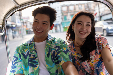 Young Asian couple tourist traveling on local Tuk Tuk Taxi exploring the city in Road in Bangkok Thailand during a holiday