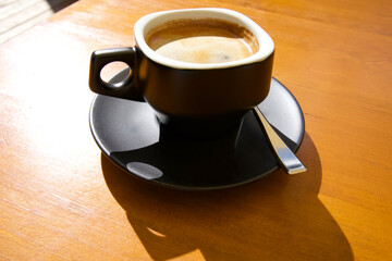 Black ceramic coffee cup with saucer and spoon on wooden table in bright sunlight creating warm ambiance