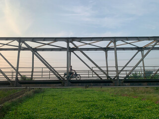 Obraz premium Silhouette of a cyclist crossing a bridge at sunset, captured from below with dramatic natural light and artistic composition.